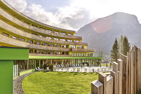 Ein modernes, halbrundes Hotelgebäude mit mehreren Balkonen steht vor einer Bergkulisse. Im Vordergrund ist ein üppiger grüner Rasen mit Sonnenliegen und einem Holzzaun unter einem bewölkten Himmel mit etwas durchbrechendem Sonnenlicht zu sehen.