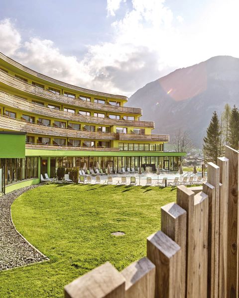 Ein modernes Hotelgebäude mit geschwungener Fassade und Balkonen blickt auf eine Rasenfläche mit Liegestühlen. Im Vordergrund verläuft ein Holzzaun, im Hintergrund sind Berge unter einem teilweise bewölkten Himmel zu sehen.