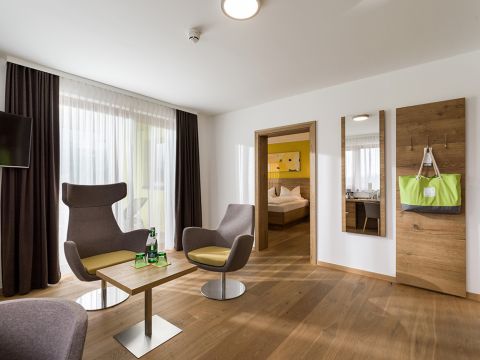 Ein modernes Hotelzimmer mit hellem Holzboden verfügt über zwei graue Stühle und einen Holztisch mit Flaschen neben einem großen Fenster mit durchsichtigen, dunklen Vorhängen. Durch eine offene Tür ist ein Bett mit weißer Bettwäsche und einer gelben Akzentwand sichtbar.