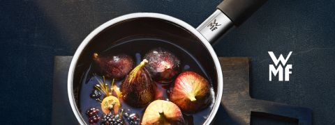 Ein Kochtopf gefüllt mit Feigen, Brombeeren, Rosmarin und Zitronenscheiben in einer dunklen Flüssigkeit. Der Topf hat einen schwarzen Griff und an der Seite die Buchstaben „WMF“. Die Szene spielt auf einer dunklen, strukturierten Oberfläche.