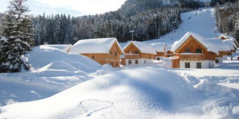Eine verschneite Landschaft mit Holzhütten inmitten von Schneewehen. Im Hintergrund sind Kiefern und hohe Hügel zu sehen. Im Vordergrund ist eine Herzform in den Schnee gezeichnet. Skifahrer fahren den Hügel hinunter.