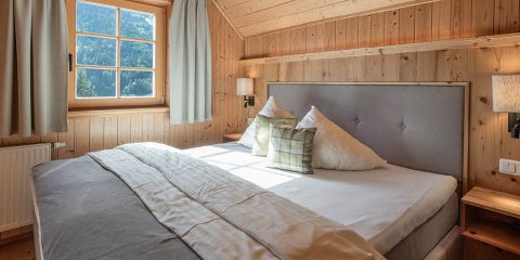 Gemütliches Schlafzimmer mit Holzwänden und -decke, mit einem großen Bett mit weißen und karierten Kissen und einem grauen Kopfteil. Sonnenlicht strömt durch ein Fenster mit hellen Vorhängen und gibt den Blick auf die malerischen Berge frei. Auf jeder Seite des Bettes befinden sich zwei Wandlampen.