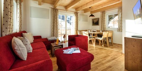 Helles Wohnzimmer mit rotem Sofa, Holzdecke und hellen Holzböden. Esstisch mit Stühlen am Fenster. Dekorative Vorhänge und gerahmte Kunstwerke an den Wänden. Natürliches Licht durchflutet den Raum durch große Fenster.