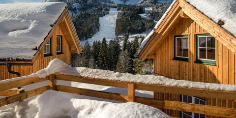 Schneebedeckte Holzhütten mit grün umrandeten Fenstern liegen eingebettet in einer Berglandschaft. Ein Holzzaun säumt den Vordergrund, während im Hintergrund dichte immergrüne Bäume und weitläufige Skipisten unter einem klaren blauen Himmel zu sehen sind.
