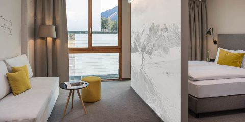 Modernes Hotelzimmer mit Sitzecke mit weißem Sofa, rundem Tisch und gelbem Ottoman. Neben der Sitzecke steht ein Bett mit grauen und gelben Akzenten. Großes Fenster mit Blick auf die Berge und ein Wandgemälde mit schneebedeckten Gipfeln an der Wand.