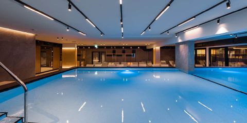 Innenpool mit ruhigem blauem Wasser und moderner Beleuchtung. Mehrere Liegestühle sind entlang einer Wand neben großen Glasfenstern angeordnet. Die Decke ist mit eleganten, parallel zueinander verlaufenden Einbauleuchten ausgestattet, die ein modernes Ambiente schaffen.