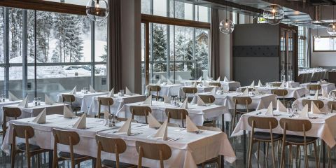 Ein geräumiges Restaurant mit großen Fenstern, die eine verschneite Landschaft draußen zeigen. Der Raum verfügt über ordentlich arrangierte Tische mit weißen Tischdecken und gefalteten weißen Servietten und Besteck. Holzstühle ergänzen das moderne Dekor.