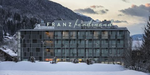Modernes Hotelgebäude mit dem Namen „Franz Ferdinand“ auf der Spitze, vor einer verschneiten Landschaft mit Bäumen und Bergen im Hintergrund. Das Gebäude verfügt über Glasbalkone und ist von Schnee umgeben.