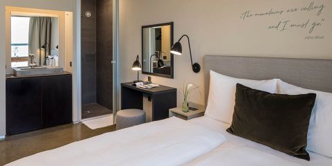 Ein modernes Hotelzimmer mit einem ordentlich gemachten Bett mit weißer Bettwäsche und grünen Kissen. Es gibt einen kleinen Schreibtisch mit einem Spiegel, eine Lampe und ein sichtbares Badezimmer. An der Wand hängt das Zitat: „Die Berge rufen und ich muss gehen.“.