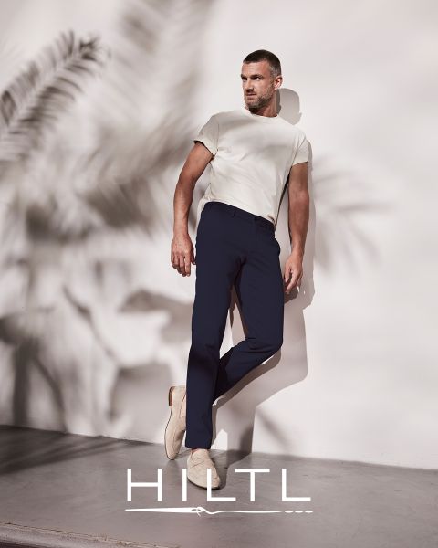 Ein Mann in weißem T-Shirt und dunkelblauer Hose lehnt an einer Wand mit Palmenblättern. Er trägt beige Schuhe. Die Szenerie ist minimalistisch. Im unteren Bildbereich steht das Wort „HILTL“.