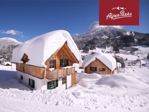Schneebedeckte Holzhütte in einer bergigen Winterlandschaft. Im Hintergrund ist eine weitere ähnliche Hütte teilweise zu sehen. Der Himmel ist klar und die Berge sind mit Schnee bedeckt. Rotes Logo in der oberen rechten Ecke mit dem Text „AlpenParks“.
