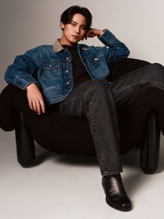 Eine Person mit dunklem Haar sitzt lässig auf einem schwarzen Stuhl. Sie trägt eine Jeansjacke über einem schwarzen Hemd, dunkle Jeans und schwarze Stiefel. Das Ambiente ist minimalistisch, mit neutralen Farben und sanfter Beleuchtung.