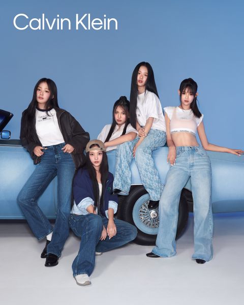 Fünf Personen posieren in blauen Jeans-Outfits vor einem blauen Auto. Sie strahlen einen lässigen, selbstbewussten Stil aus. Oben links im Bild steht in weißer Schrift „Calvin Klein“. Der Hintergrund ist einfarbig blau.