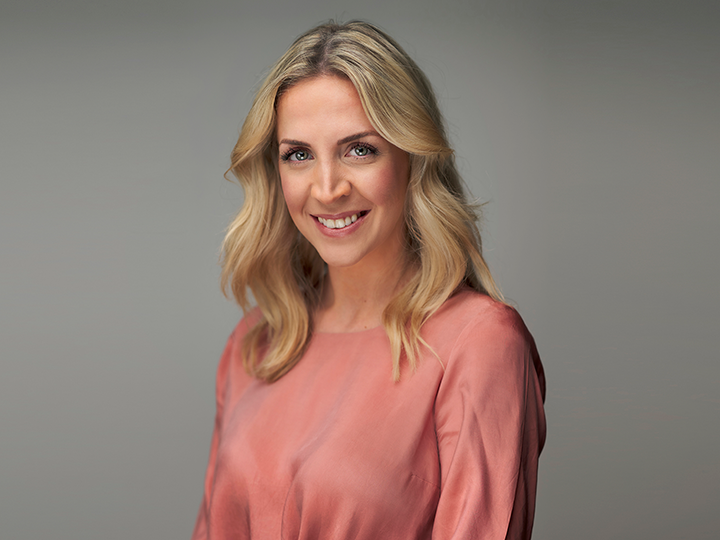 Eine Person mit langen, welligen blonden Haaren und einem warmen Lächeln trägt ein langärmliges, korallenfarbenes Oberteil. Der Hintergrund ist in einem neutralen Grau gehalten, was dem Bild eine professionelle und freundliche Ausstrahlung verleiht.