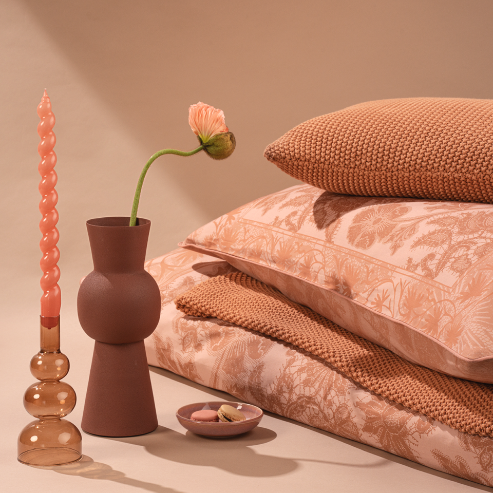 Gemütliches Stillleben mit gefalteten gestrickten und gemusterten Kissen in sanften Terrakottatönen. Eine gewellte rosa Kerze in einem Glashalter, eine einzelne Mohnblume in einer Keramikvase und eine kleine Schale mit Makronen sorgen für eine warme, einladende Note.