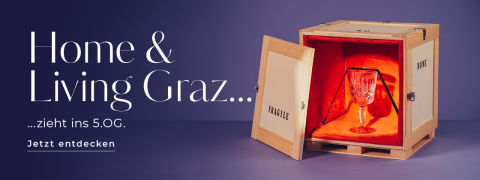 Eine Holzkiste mit der Aufschrift „Fragile“ ist geöffnet und gibt den Blick frei auf eine rote Samtfüllung und einen Glaspokal. Auf violettem Untergrund steht „Home & Living Graz zieht ins 5. OG. Jetzt entdecken“.