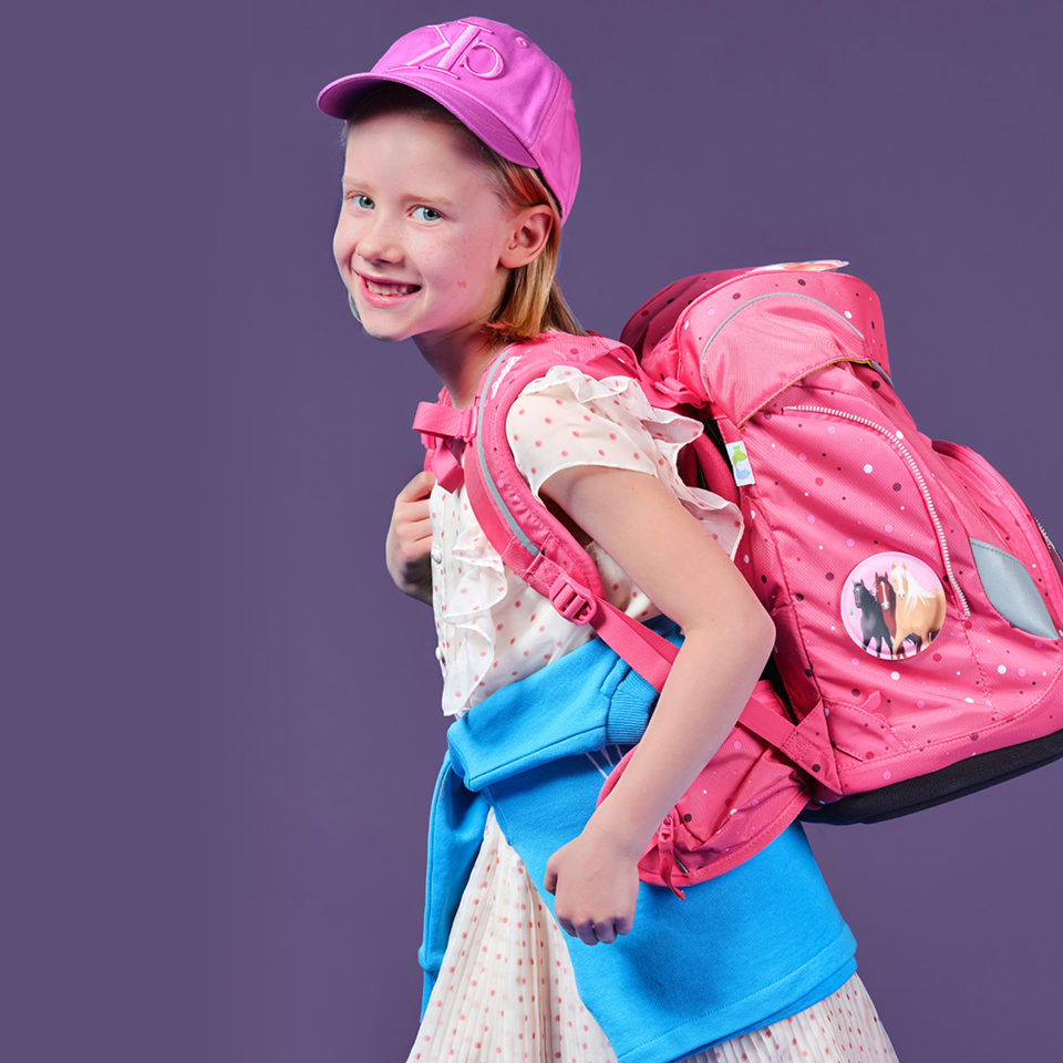 Junges Mädchen mit rosa Mütze, gepunktetem Kleid und blauem Pullover um die Taille. Sie lächelt und trägt einen großen rosa Rucksack mit einem Pferdeknopf vor einem violetten Hintergrund.