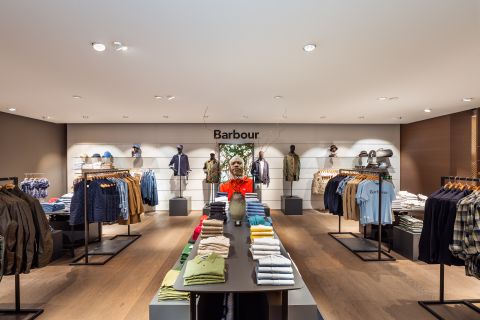 Inneneinrichtung eines Bekleidungsgeschäfts mit ordentlich angeordneten Regalen und Ablagen, auf denen verschiedene Hemden, Jacken und Accessoires ausgestellt sind. Auf einem Tisch in der Mitte liegen gefaltete Kleidungsstücke. Das Geschäft hat eine warme, einladende Atmosphäre mit natürlichem Holzboden.