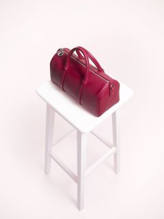 Eine rote Lederhandtasche mit zylindrischer Form steht auf einem weißen Hocker vor einem schlichten, neutralen Hintergrund. Die Tasche hat abgerundete Griffe und metallische Akzente.