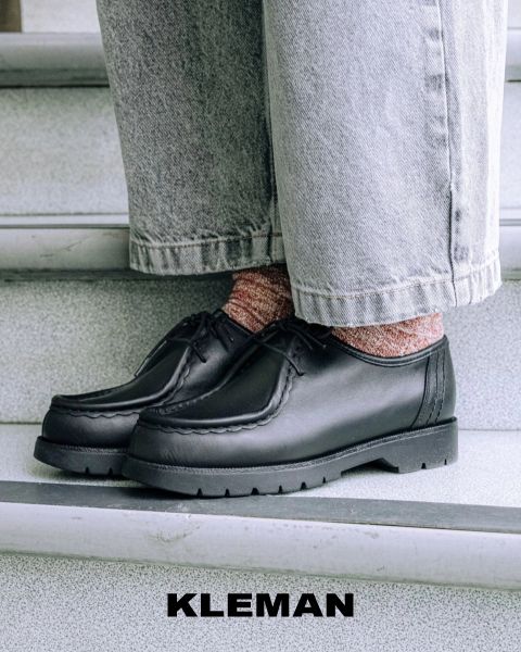 Nahaufnahme einer Person in schwarzen Lederschuhen mit rosa gemusterten Socken und hellen, weit geschnittenen Jeans, die auf einer grauen Treppe steht. Der Markenname "KLEMAN" erscheint am unteren Rand des Bildes.