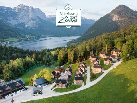 Luftaufnahme von Narzissendorf Zloam, einem malerischen Dorf mit Holzhäusern, eingebettet in üppige grüne Hügel. Ein ruhiger See und Berge bilden eine atemberaubende Kulisse. Oben in der Mitte befindet sich ein Logo für Narzissendorf Zloam.