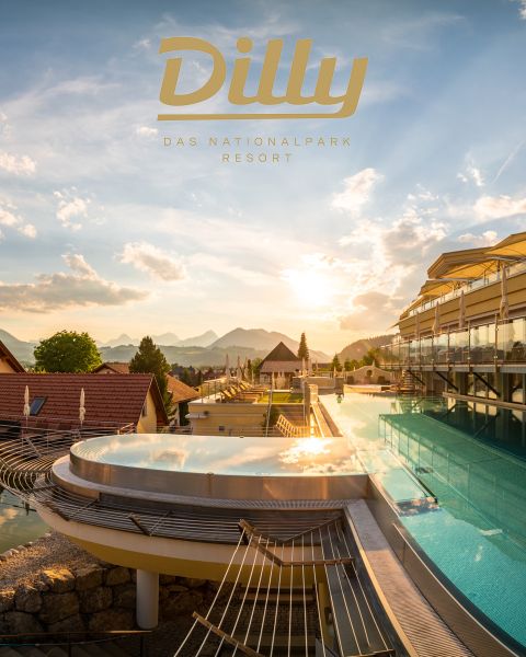 Poolterrasse auf dem Dach bei Sonnenuntergang mit Blick auf die Berge. Überlappende runde Pools sind von Glasgeländern umgeben. Das Gebäude auf der rechten Seite hat große Fenster. Der Text oben lautet „Dilly Das Nationalpark Resort“.