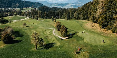 Luftaufnahme eines üppigen, grünen Golfplatzes, umgeben von Bäumen und Hügeln. Mehrere Golfer sind auf dem Platz und Golfwagen stehen in der Nähe. Die Landschaft umfasst Sandbunker und eine malerische Kulisse mit entfernten Bergen.