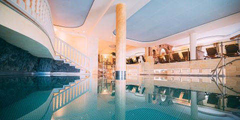 Innenpoolbereich mit ruhigem Wasser, in dem sich eine Säule und die umgebende Architektur spiegeln. Eine Treppe auf der linken Seite führt nach oben, während auf der rechten Seite Reihen von Liegestühlen stehen. Die Atmosphäre ist ruhig und luxuriös mit sanfter Beleuchtung.