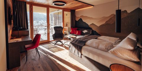 Ein gemütliches Hotelzimmer verfügt über ein großes Bett mit weißen Kissen und einer grauen Decke, einen Schreibtisch mit rotem Stuhl und einen gepolsterten Sessel. Das Zimmer hat Holzböden und einen Balkon mit malerischem Bergblick. Warme Beleuchtung schafft eine einladende Atmosphäre.
