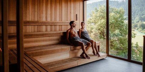 Ein Mann und eine Frau sitzen in Handtücher gehüllt in einer hölzernen Sauna. Sie sitzen auf Holzbänken, schauen einander an und lächeln. Große Fenster geben den Blick frei auf Bäume und sanfte Hügel unter einem klaren Himmel.