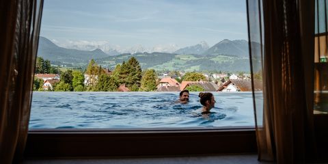 Zwei Menschen schwimmen in einem Infinity-Pool mit malerischer Aussicht auf üppig grüne Hügel und Berge in der Ferne unter einem klaren blauen Himmel, eingerahmt durch offene Vorhänge aus dem Inneren eines Gebäudes.