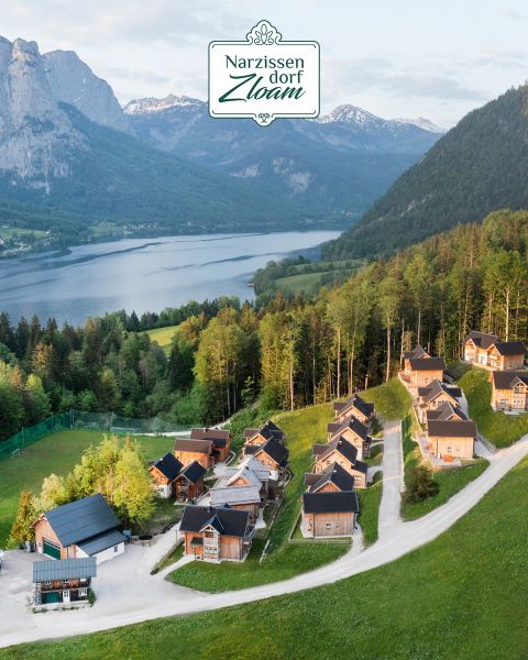 Luftaufnahme eines ruhigen Dorfes mit Holzhütten inmitten üppiger Vegetation und einem See, umgeben von Bergen. Der Dorfname „Narzissendorf Zloam“ wird oben im Bild angezeigt.