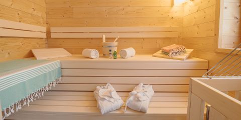 Ein Sauna-Innenraum aus Holz mit zwei aufgerollten Handtüchern und einem Eimer mit Schöpfkelle auf der Bank. Hausschuhe liegen auf den Dielen. Ein blaues Handtuch und gefaltete Bademäntel verleihen der natürlichen Holzausstattung einen Hauch von Farbe.