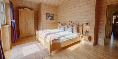 Ein gemütliches Schlafzimmer aus Holz mit einem Doppelbett mit rosa Kissen und einer passenden Decke. Es gibt einen hölzernen Kleiderschrank, eine kleine Vase mit Blumen und ein gerahmtes Bild an der Wand. Sonnenlicht fällt durch das Fenster auf einen gestreiften Teppich.