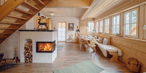 Gemütliche Inneneinrichtung einer Holzhütte mit Kamin, Essbereich und großen Fenstern. Brennholz wird neben dem Kamin gelagert und Holzmöbel schmücken den Raum. Eine Treppe führt in die obere Etage und Licht strömt durch die Fenster.