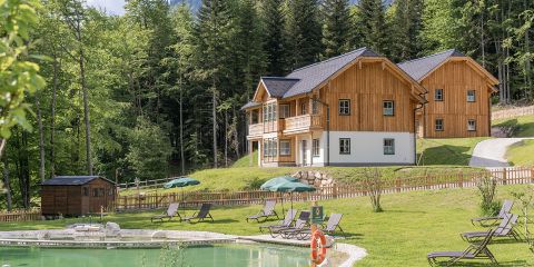 Zwei Holzhütten mit Balkon liegen eingebettet in einer üppigen Waldlandschaft. Im Vordergrund ist ein ruhiger Teich von Liegestühlen und Sonnenschirmen umgeben. Die Szene ist friedlich und idyllisch, mit lebendigem Grün und klarem Himmel.
