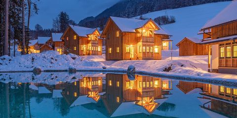 Eine Winterlandschaft mit Holzhütten, die von warmem Licht beleuchtet und von Schnee umgeben sind. Die Gebäude spiegeln sich in einem ruhigen Gewässer im Vordergrund, im Hintergrund sind ein Wald und ein Berg unter einem dämmrigen Himmel zu sehen.
