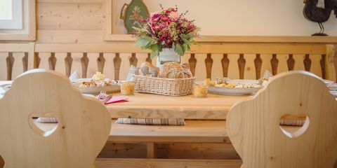 Ein gemütlicher Esstisch, der für eine Mahlzeit gedeckt ist, mit Holzstühlen mit herzförmigen Ausschnitten. In der Mitte stehen ein Weidenkorb mit Brot und eine Vase mit bunten Blumen. Auf dem Tisch sind Teller mit Essen und Gläser angeordnet. Die Umgebung ist rustikal und einladend.
