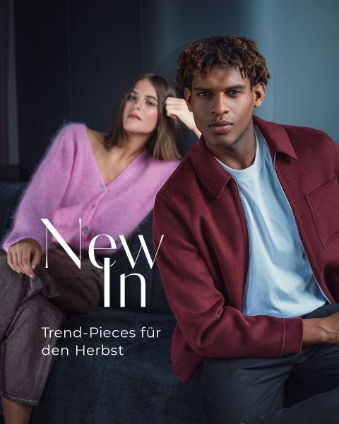 Eine Frau in einem rosa Pullover und ein Mann in einer burgunderroten Jacke posieren in einem Innenraum. Der Text lautet: „Neu eingetroffen. Trendteile für den Herbst.“.