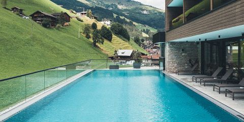 Ein moderner Außenpool mit Glasgeländer bietet Aussicht auf einen üppigen, grünen Hügel mit Holzhäusern. Liegestühle säumen den Pool und im Hintergrund ist eine Berglandschaft unter einem bewölkten Himmel zu sehen.