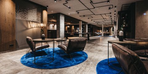 Geräumige, moderne Lobby mit gedämpfter Beleuchtung, Lederstühlen und runden Tischen auf leuchtend blauen Teppichen. An der Decke sind freiliegende Rohrleitungen angebracht und die Wände sind eine Mischung aus Holzvertäfelung und strukturierten Oberflächen. Minimalistisches Dekor verleiht dem Ganzen eine elegante Note.