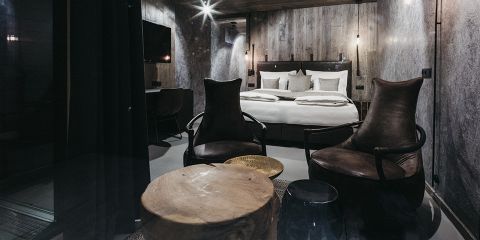 Ein gemütliches, schwach beleuchtetes Hotelzimmer mit einem großen Bett, einem hölzernen Kopfteil und moderner Einrichtung. Es gibt zwei dunkle Ledersessel gegenüber einem runden hölzernen Couchtisch und einem an der Wand montierten Fernseher. Die Wände und die Decke haben ein rustikales, holzgetäfeltes Design.