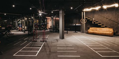 Geräumiges, modernes Fitnessstudio mit gedämpfter Beleuchtung und verschiedenen Trainingsgeräten wie Gewichten und Ringen. Neben der Treppe steht ein braunes Ledersofa und weiße Markierungen auf dem Boden kennzeichnen die Trainingsbereiche.