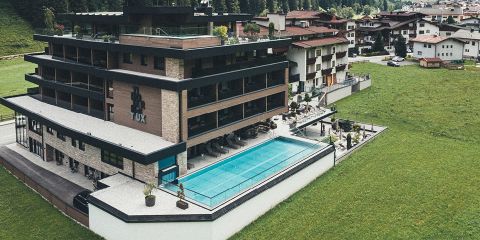 Ein modernes Hotel mit mehreren Etagen, einer Dachterrasse und einem Außenpool ist von üppigen grünen Rasenflächen umgeben. In der Nähe befinden sich traditionelle Häuser mit schrägen Dächern. Die Umgebung ist malerisch und ruhig.