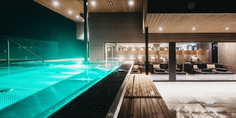 Moderner Außenpool mit türkis leuchtendem Wasser bei Nacht. Angrenzende überdachte Lounge mit großen Glasfenstern, holzgetäfelter Decke und gepolsterten Sitzgelegenheiten. Der beleuchtete Raum schafft eine gemütliche, elegante Atmosphäre.