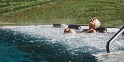Eine Person mit kurzen blonden Haaren entspannt sich in einem sprudelnden Außenpool und lehnt sich mit geschlossenen Augen zurück. Der Pool blickt auf einen grasbewachsenen Hügel und das Sonnenlicht wirft einen warmen Schein auf die Szene.