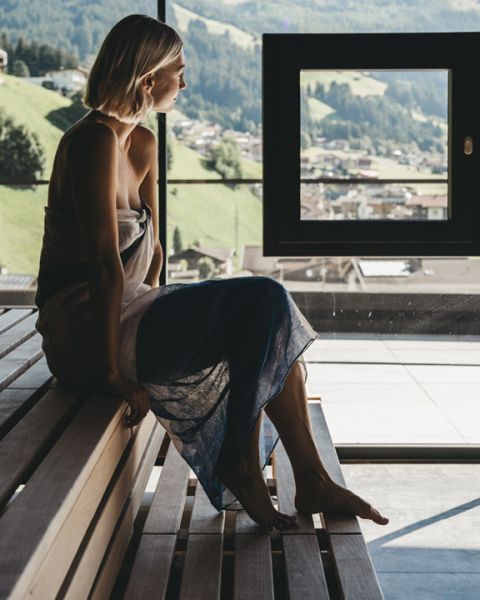 Eine Frau sitzt in ein Handtuch gehüllt auf einer Holzbank und blickt aus einem großen Fenster. Der Blick nach draußen zeigt eine malerische Hügellandschaft und eine entfernte Stadt unter einem klaren Himmel. Natürliches Licht erfüllt den Raum.