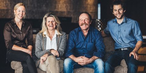 Vier Personen sitzen lächelnd auf einem Sofa. Die Gruppe besteht aus zwei Frauen links und zwei Männern rechts, alle leger gekleidet. Der Hintergrund wirkt dunkel und unscharf, was auf eine Innenaufnahme schließen lässt.
