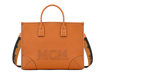 Orangefarbene MCM-Handtasche mit zwei Griffen und einem abnehmbaren schwarzen Riemen mit dem Markennamen. Die Tasche hat ein strukturiertes Design mit sichtbaren Nähten und Eckdetails und trägt auf der Vorderseite deutlich die Prägung „MCM“.