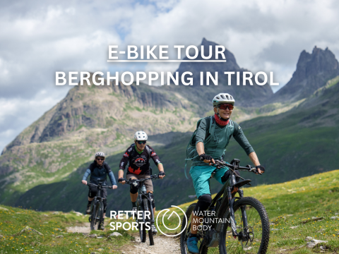 Drei Personen fahren mit E-Bikes auf einem malerischen Bergpfad in Tirol unter einem bewölkten Himmel. Der Text lautet oben „E-Bike-Tour Berghopping in Tirol“, darunter die Logos „Retter Sports“ und „Water Mountain Body“. Das Gelände ist grasbewachsen und rau.